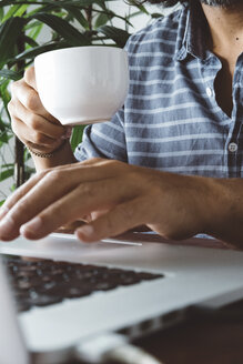 Mittelteil eines Mannes, der eine Kaffeetasse hält, während er einen Laptop-Computer auf einem Tisch zu Hause benutzt - CAVF50352