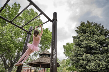 Rückansicht eines Mädchens in einem Ballettkostüm, das an einem Klettergerüst hängt, gegen einen bewölkten Himmel auf einem Spielplatz - CAVF50388