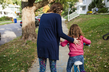 Rückansicht einer Mutter, die ihrer Tochter beim Fahrradfahren auf einem Fußweg im Herbst hilft - CAVF50419
