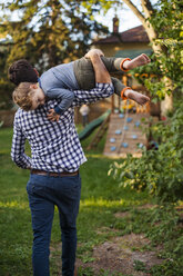 Rear view of playful father carrying son while walking on grassy field in yard during sunset - CAVF50437