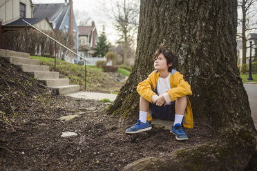 Thoughtful boy looking away while sitting by tree trunk - CAVF50439