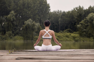 Rear view of woman meditating while sitting on pier by lake against trees during sunset - CAVF50464