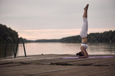 Woman practicing headstand on pier by lake against cloudy sky during sunset - CAVF50465