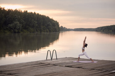 Woman practicing warrior 1 pose on pier by lake against cloudy sky during sunset - CAVF50466