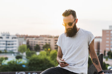 Cool bearded young man wearing sunglasses checking cell phone - KKAF02503