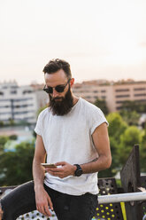 Cooler bärtiger junger Mann mit Sonnenbrille, der sein Mobiltelefon überprüft - KKAF02504