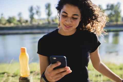 Sportliche junge Frau mit Mobiltelefon bei einer Pause am Flussufer, lizenzfreies Stockfoto