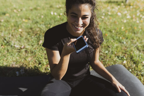 Fröhliche sportliche junge Frau, die eine Pause auf einer Wiese macht und dabei ihr Handy benutzt - FMOF00396