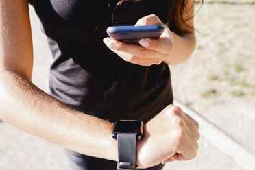 Nahaufnahme einer sportlichen jungen Frau, die ein Mobiltelefon und eine Smartwatch benutzt - FMOF00410