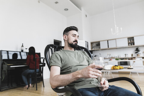 Ehepaar zu Hause, Mann trinkt Wein, Frau spielt im Hintergrund Klavier, lizenzfreies Stockfoto