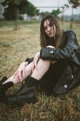 Portrait of woman in raincoat sitting on grassy field at park during rainy season - CAVF50468