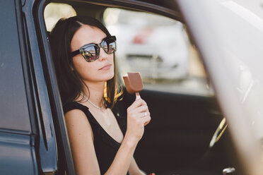 Junge Frau mit Sonnenbrille hält gefrorene Süßigkeiten in der Hand, während sie im Auto sitzt, gesehen durch ein Fenster - CAVF50491