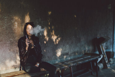 Portrait of confident young woman smoking electronic cigarette while sitting on bench against wall on footpath - CAVF50492