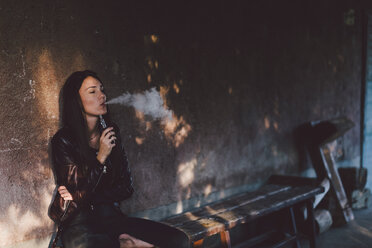 Junge Frau, die eine elektronische Zigarette raucht, während sie auf einer Bank an der Wand eines Fußwegs sitzt - CAVF50493