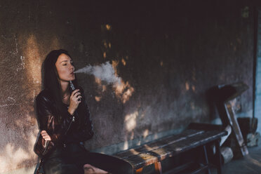 Young woman smoking electronic cigarette while sitting on bench against wall on footpath - CAVF50493