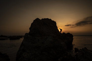 Silhouette child climbing on rock at beach against sky during sunset - CAVF50497