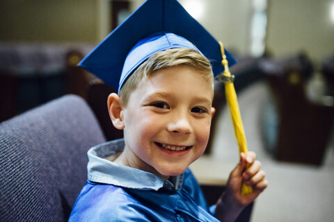 Porträt eines glücklichen Jungen im Abschlusskleid auf einem Stuhl sitzend, lizenzfreies Stockfoto