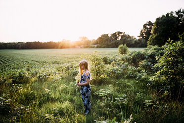 Seitenansicht eines Mädchens, das Blumen hält, während es auf einem grasbewachsenen Feld vor einem klaren Himmel bei Sonnenuntergang steht - CAVF50526