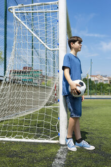 Seitenansicht eines Jungen, der einen Fußball hält, während er am Netz auf einer Wiese vor dem Himmel steht - CAVF50549