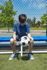 Junge hält Fußball, während er auf einer Bank am Zaun des Unterstandes sitzt - CAVF50553