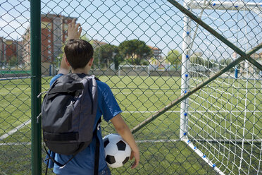 Rückansicht eines Jungen mit Rucksack, der einen Fußball hält, während er am Zaun des Unterstandes steht - CAVF50554