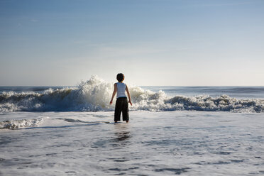 Rear view of boy playing with waves while standing in sea against sky during sunset - CAVF50566