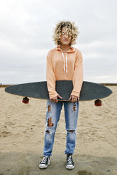 Junge Frau mit lockigem blondem Haar, rosa Kapuzenpulli und zerrissenen Jeans, steht am Sandstrand, hält ein Skateboard und schaut in die Kamera. - MINF09088