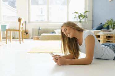 Smiling young woman lying on floor in her loft looking at cell phone - KNSF04994