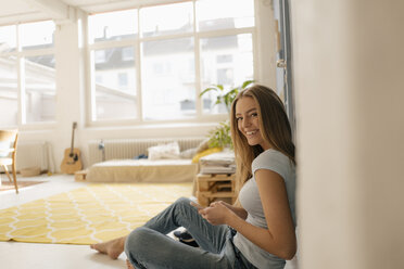 Portrait of smiling young woman sitting on the floor in her loft using smartphone - KNSF05012