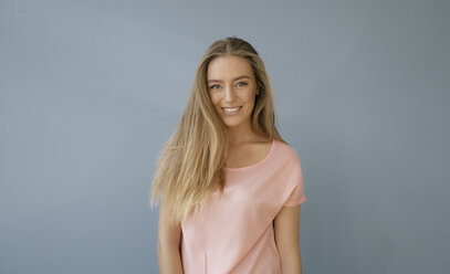Porträt einer lächelnden jungen Frau mit rosa T-Shirt vor grauem Hintergrund - KNSF05017