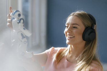 Porträt einer lächelnden jungen Frau mit Kopfhörern, die einen Spielzeugroboter betrachtet - KNSF05020