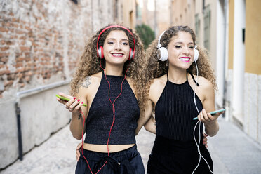 Porträt von lächelnden Zwillingsschwestern, die mit Kopfhörern und Mobiltelefonen in der Stadt Musik hören - GIOF04722