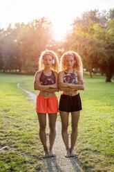 Portrait of twin sisters standing side by side in a park listening music with headphones - GIOF04754