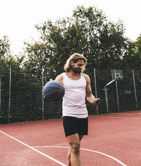 Junger Mann verlässt den Basketballplatz, trägt einen Ball und benutzt ein Smartphone - UUF15568