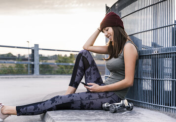 Young woman sitting on ground after dumbbell training, using smartphone - UUF15587
