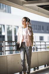 Young businesswoman walking in parking garage, listeing music with her headphones - UUF15607