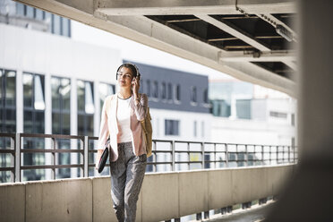 Young businesswoman walking in parking garage, listeing music with her headphones - UUF15609