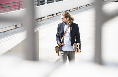 Young businessman carrying skateboard, using smartphone - UUF15620