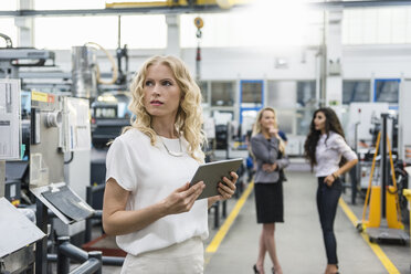 Frau hält Tablet in Fabrikhalle mit zwei Frauen im Hintergrund - DIGF05285