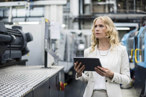 Frau mit Tablet an einer Maschine in einer Fabrikhalle und schaut sich um, lizenzfreies Stockfoto
