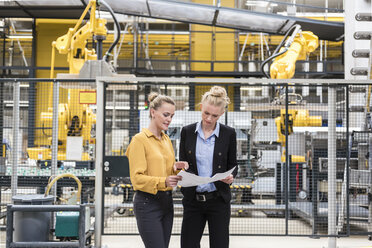 Two women discussing plan in factory shop floor with industrial robot - DIGF05388