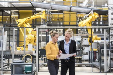 Two women discussing plan in factory shop floor with industrial robot - DIGF05389