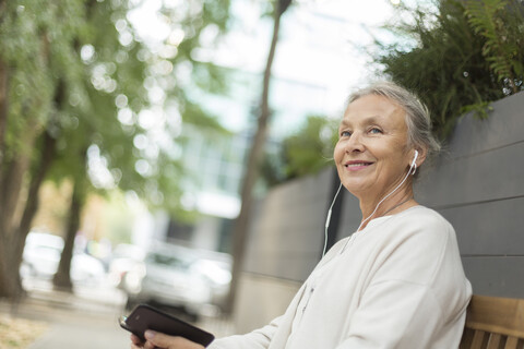 Lächelnde ältere Frau sitzt auf einer Bank im Freien mit Handy und Kopfhörern, lizenzfreies Stockfoto