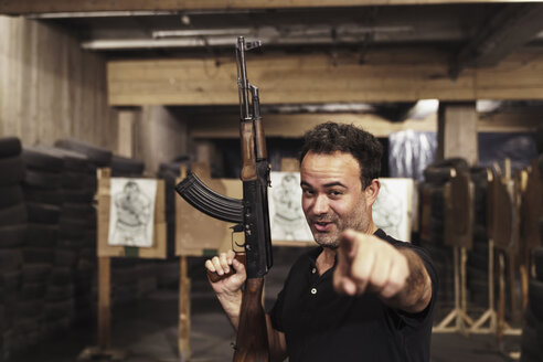 Porträt eines Mannes mit einem Gewehr, der mit dem Finger auf eine Schießhalle zeigt - KKAF02596