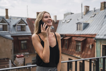 Lächelnde blonde Frau am Telefon, die auf einem Balkon steht und nach oben schaut - KKAF02617