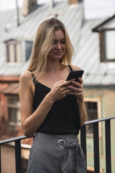 Porträt einer lächelnden blonden Frau, die auf einem Balkon steht und auf ihr Handy schaut - KKAF02621