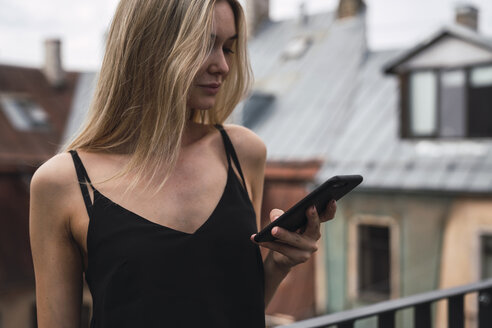 Blonde Frau auf dem Balkon schaut auf ihr Handy - KKAF02622