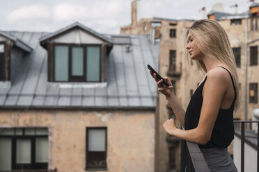 Blonde junge Frau steht auf einem Balkon und schaut auf ihr Handy - KKAF02623