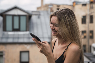 Lächelnde blonde junge Frau auf der Dachterrasse, die auf ihr Handy schaut - KKAF02625