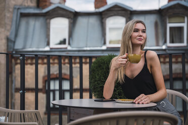 Porträt einer blonden jungen Frau, die auf einer Dachterrasse sitzt und eine Tasse Kaffee trinkt - KKAF02629